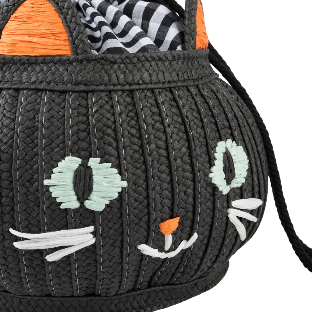 A black woven handbag with a cat face design, orange ears, and green eyes. It features a black and white checkered fabric peeking out from the top.
