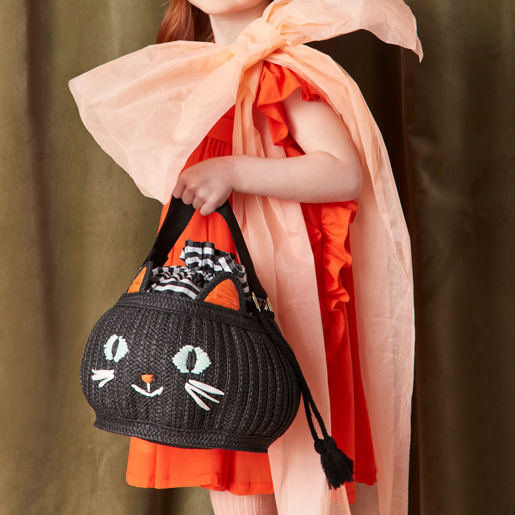 A child in a bright orange dress with a large, light pink bow holds a black woven handbag shaped like a cat, featuring playful details.