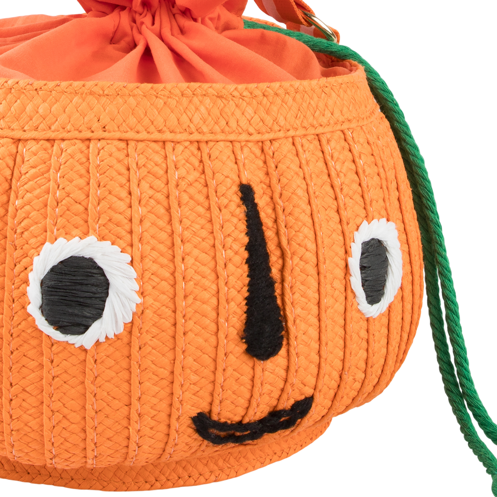 Orange pumpkin-shaped bag with a smiling face, featuring bold black stitched eyes and mouth. It has a drawstring top and green tassel detail. Cheerful tone.