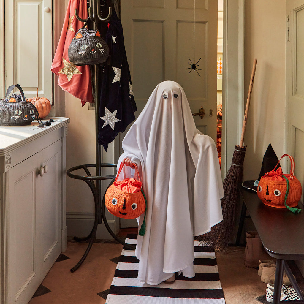 A child dressed as a ghost, with a white sheet and googly eyes, holds a jack-o'-lantern basket. The cozy room has Halloween decor like pumpkins, a broom, and starry cloaks.
