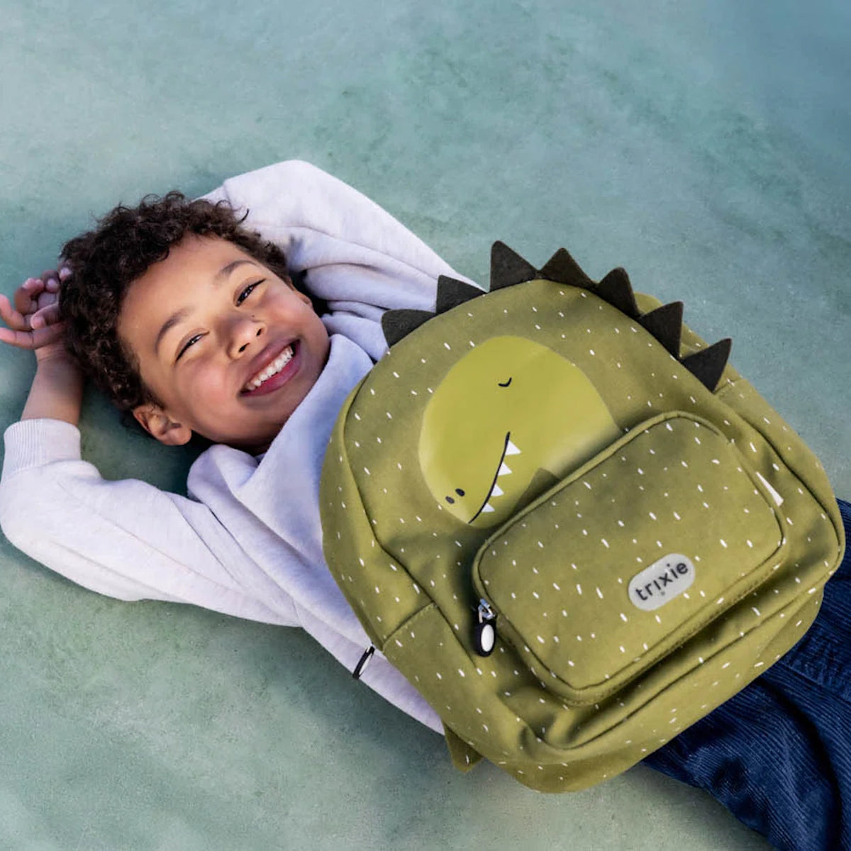 A smiling boy lying down with a green, speckled dinosaur backpack featuring spikes and a front pocket.