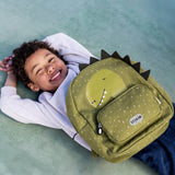 A smiling boy lying down with a green, speckled dinosaur backpack featuring spikes and a front pocket.