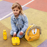 A young child in a denim jacket sits outdoors next to a yellow lion backpack, lunch box, and water bottle.