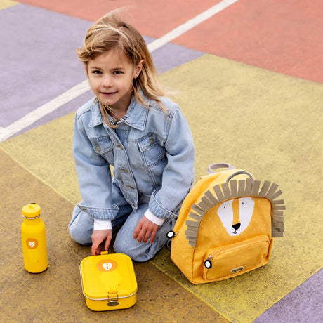A young child in a denim jacket sits outdoors next to a yellow lion backpack, lunch box, and water bottle.