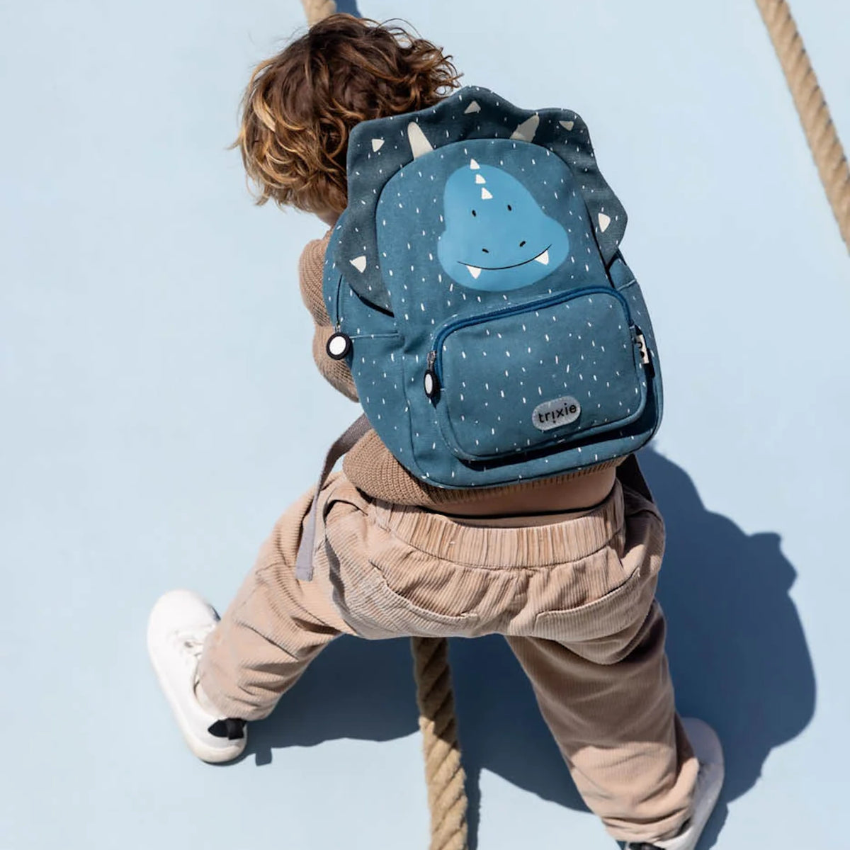 A child climbing a structure while wearing a blue triceratops dinosaur backpack with white speckles.