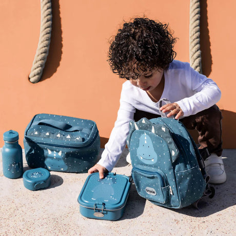 A young child handles a matching set of blue triceratops gear: a backpack, lunch box, bag, and water bottle.