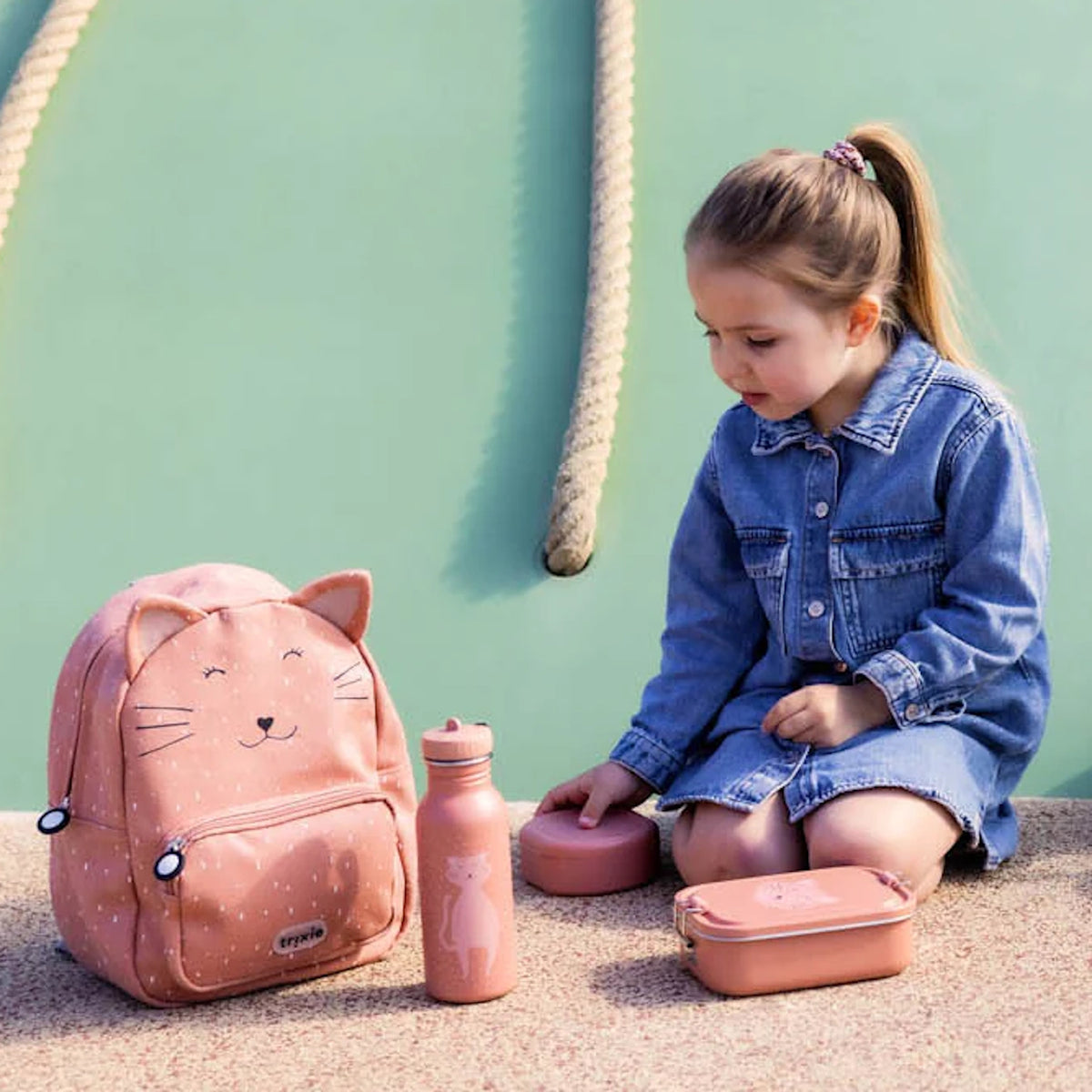 A young girl in a denim shirt next to a pink cat-themed backpack, water bottle, and lunch container.