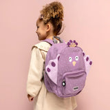 A young girl from the side wearing a purple peacock backpack with white speckles against a pink background.
