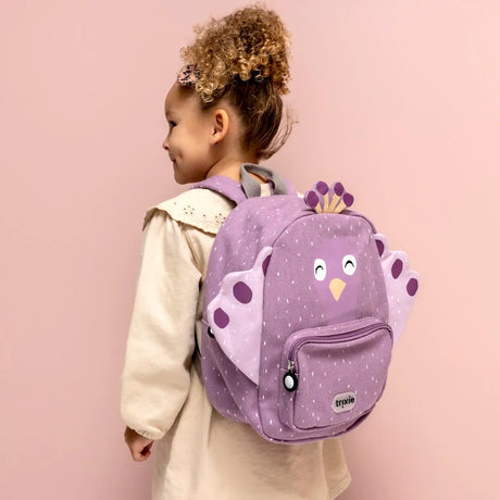 A young girl from the side wearing a purple peacock backpack with white speckles against a pink background.