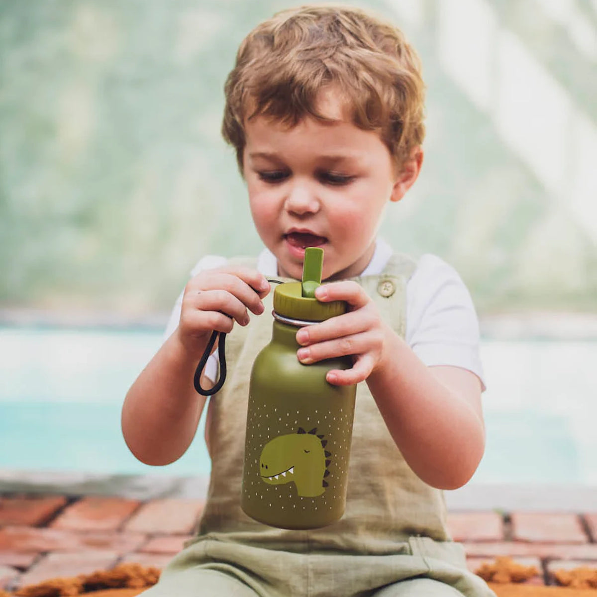 A young boy in overalls sits outdoors, holding a green kids water bottle with a cartoon dinosaur graphic.