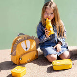 Young girl drinking from a yellow water bottle next to a matching lion backpack and lunch boxes.