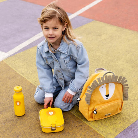 Young girl in a denim outfit sitting on a colorful ground with a yellow lion backpack, lunchbox, and bottle.
