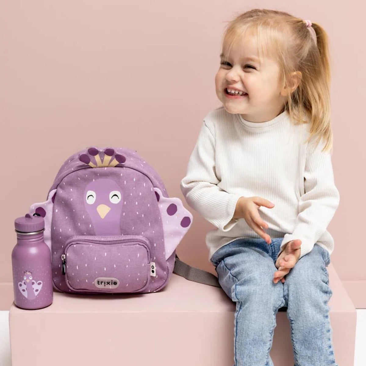 Smiling young girl sitting next to a matching purple peacock backpack and water bottle.