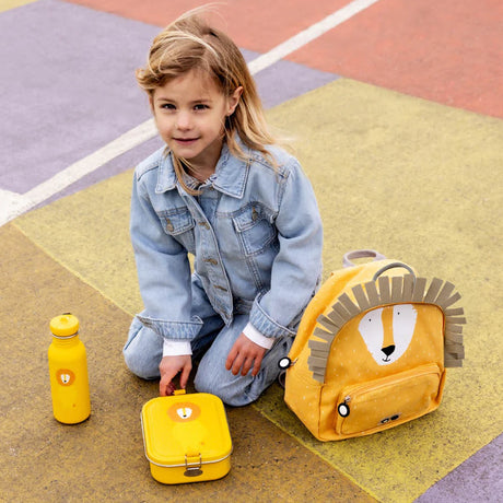 A young girl in denim sitting outdoors with a matching yellow lion-themed backpack, lunch box, and water bottle.