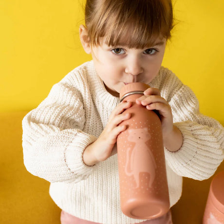 A young girl in a knit sweater drinking from a pink cartoon cat water bottle against a yellow background.