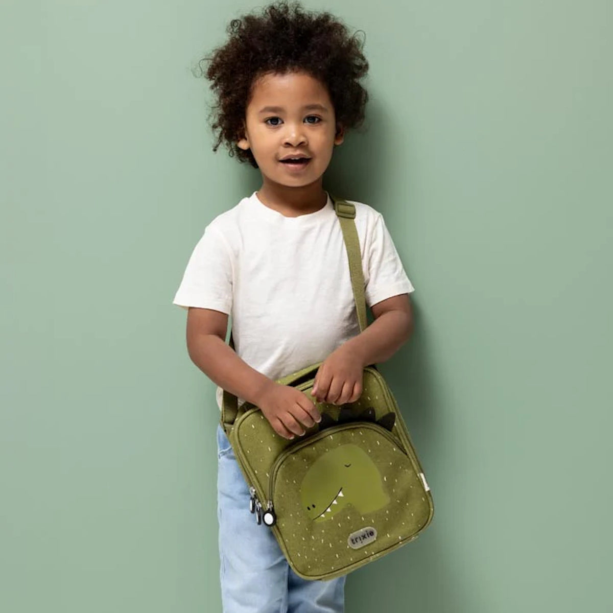 A young child standing and holding a green dinosaur-themed lunch bag with a shoulder strap against a green wall.