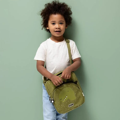 A young child standing and holding a green dinosaur-themed lunch bag with a shoulder strap against a green wall.