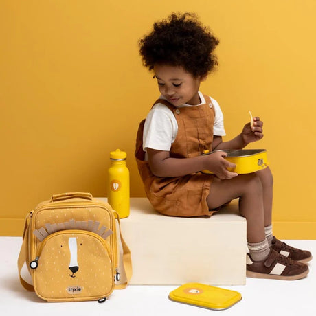Young child sitting next to a yellow lion-themed lunch bag and water bottle while holding a lunch box.