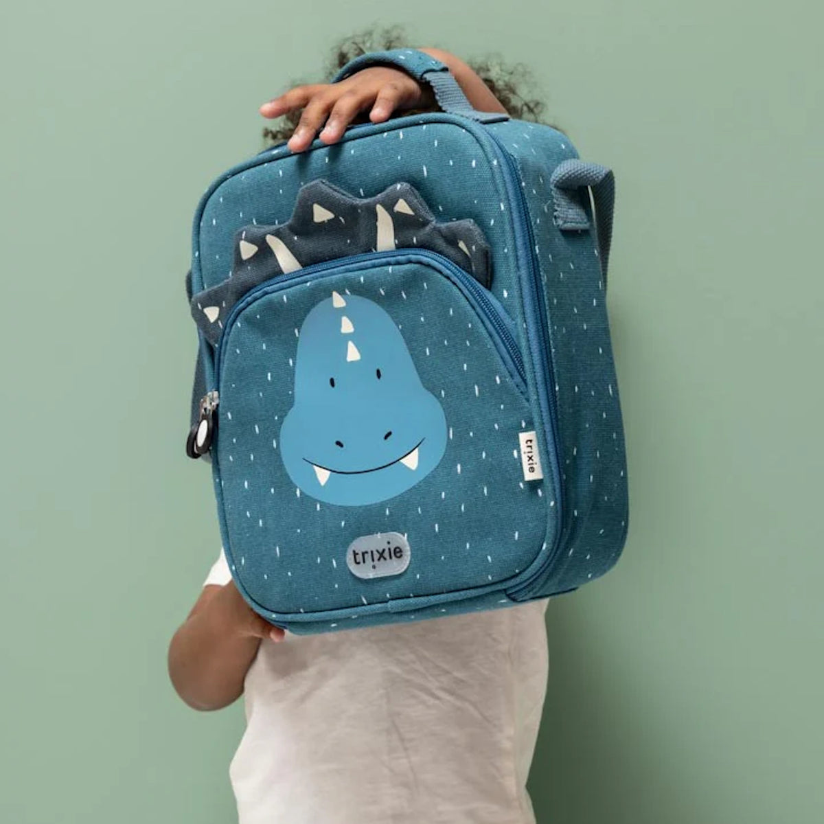 A child holds up a teal dinosaur lunch bag by its handle against a solid green background.