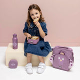 A young girl in a denim dress eats a sandwich next to a purple peacock themed lunch bag and water bottle.