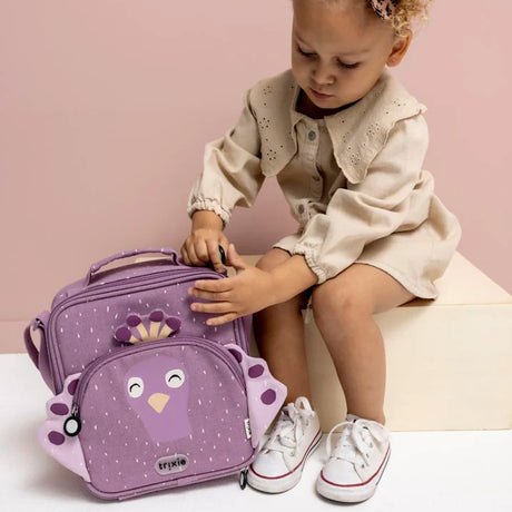 A young child sitting on a wooden block opens a purple peacock-themed lunch bag against a pink background.