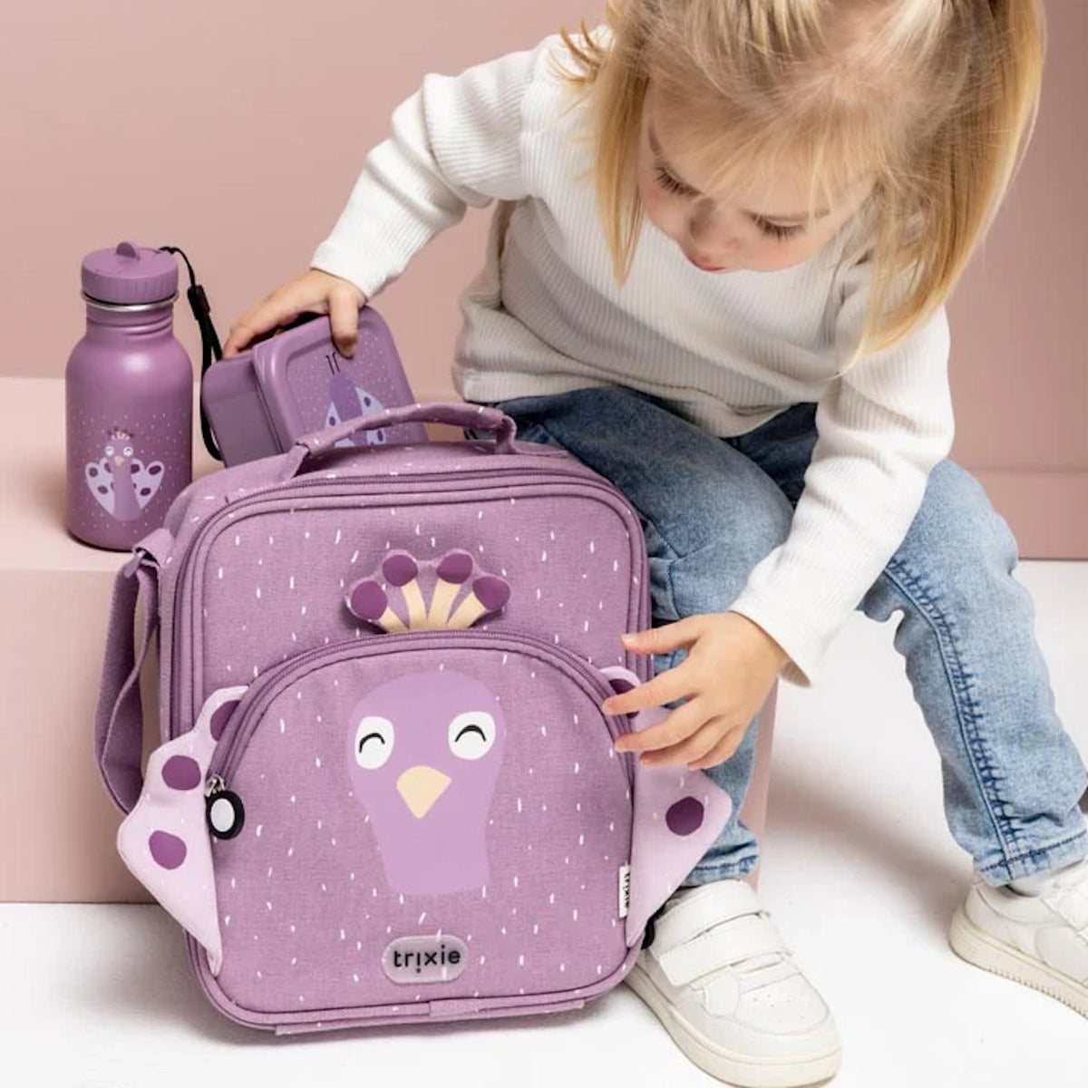 A young child sitting next to a purple peacock lunch bag, water bottle, and snack box.