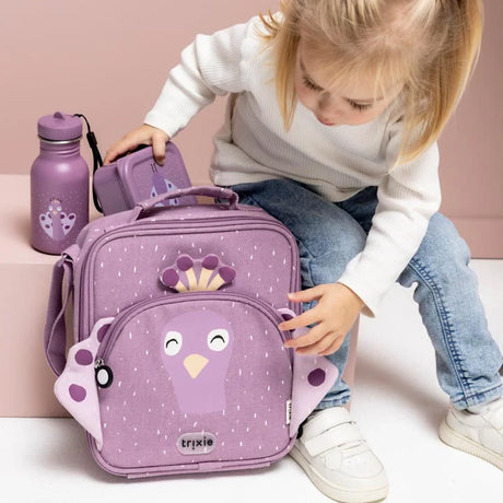 A young child sitting next to a purple peacock lunch bag, water bottle, and snack box.