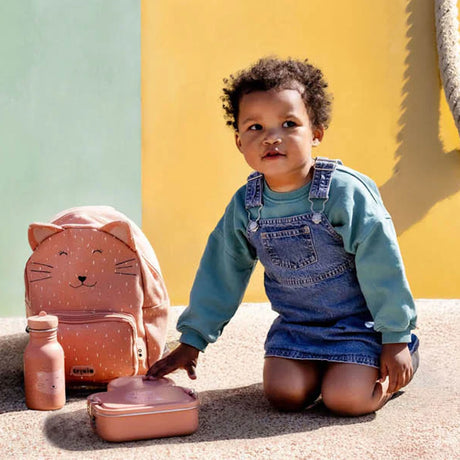A young child in denim overalls kneeling next to a pink Trixie cat backpack, lunch box, and water bottle.