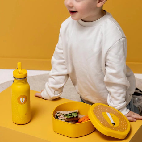 A young child sitting by an open, yellow lion-shaped lunch box containing a wrap next to a matching water bottle.