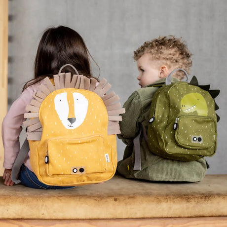Two toddlers from behind wearing patterned animal backpacks: a yellow lion and a green dinosaur.