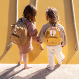 Two young children from behind holding hands while wearing small animal backpacks: a brown dog and a yellow lion.