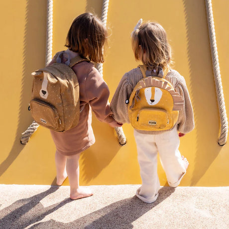 Two young children from behind holding hands while wearing small animal backpacks: a brown dog and a yellow lion.