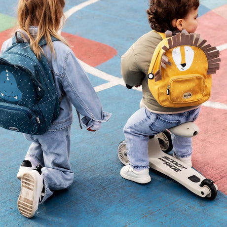 Two young children playing outside, wearing a blue dinosaur backpack and a yellow lion backpack.