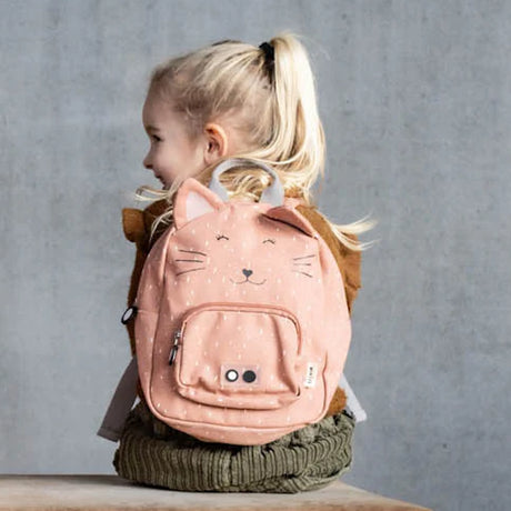 A young girl with blonde hair from behind, wearing a pink cat-themed backpack against a gray wall.