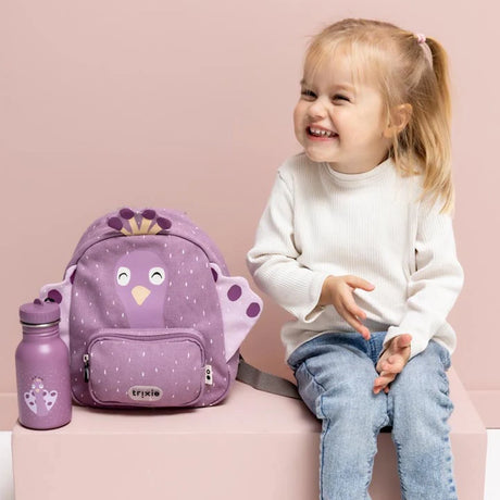 A smiling young girl sits next to a purple peacock-themed kids backpack and matching water bottle.