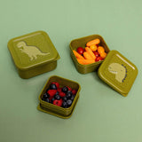 Three green dinosaur-themed nesting snack boxes, filled with berries and baby carrots, on a green background.