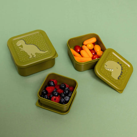 Three green dinosaur-themed nesting snack boxes, filled with berries and baby carrots, on a green background.
