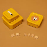 Three yellow lion-themed nesting snack boxes displayed with alphabet crackers on a solid ochre background.