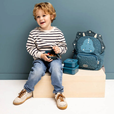 A young boy sitting with blue dinosaur nesting snack boxes and a matching backpack against a blue wall.