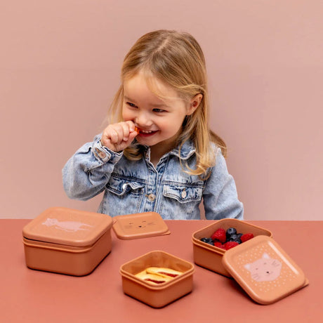 Smiling child eating a snack next to three pink cat-themed nesting boxes, two filled with fresh berries and food.