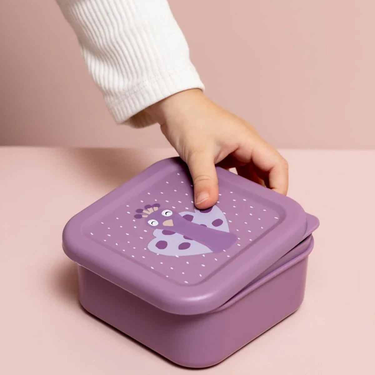 A child's hand opening a purple nesting snack box with a cartoon peacock illustration on the lid.