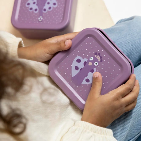 A child holding a purple square snack box with a cartoon peacock illustration on the lid.