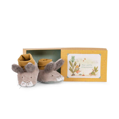 Plush grey rabbit baby slippers with mustard yellow cuffs next to their yellow illustrated gift box.