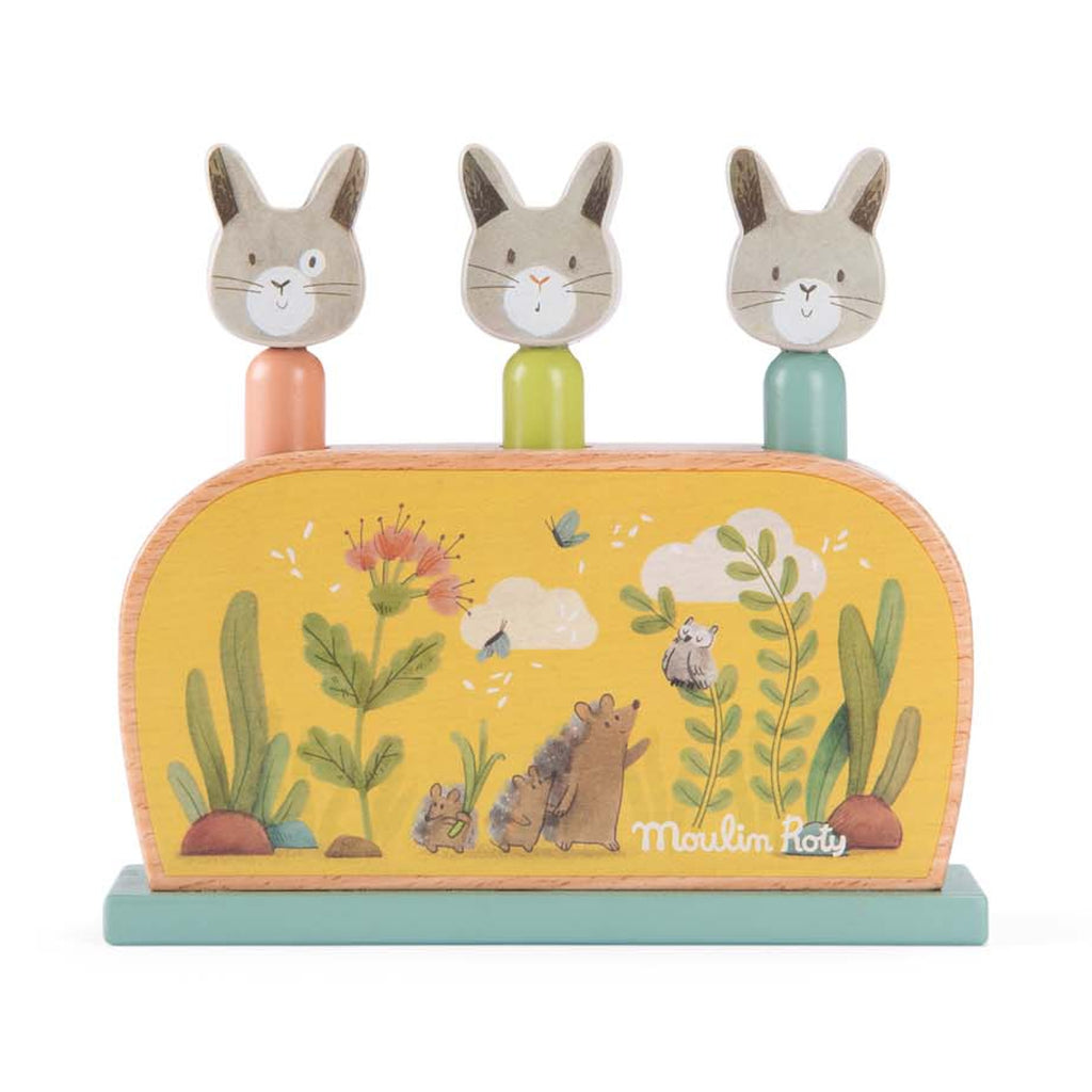 Wooden toy with three smiling rabbit heads on colored pegs atop a base. Yellow backdrop features whimsical plant and animal illustrations. Bright, playful.