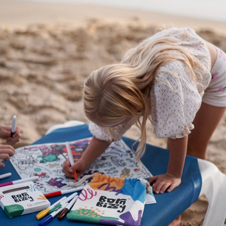 A child with blonde hair colors a mat on a beach chair, with markers and a "Born Bizzy" pouch nearby.
