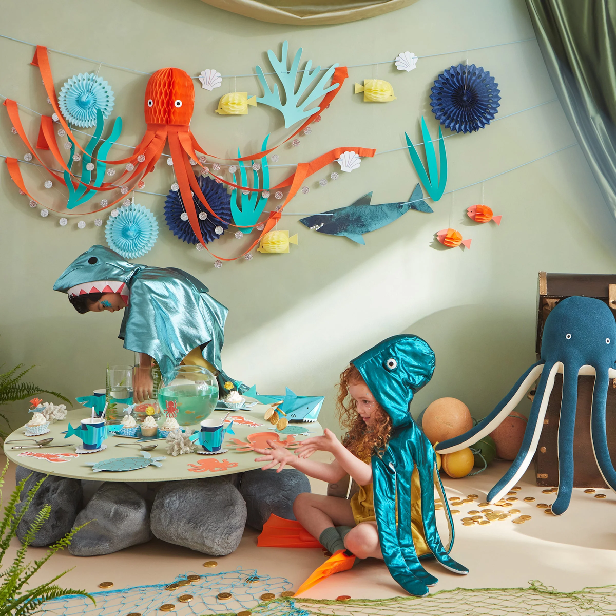 Children in ocean costumes playing near a decorated party table under an Under the Sea themed garland.