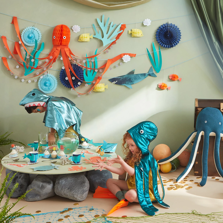 Children in ocean costumes playing near a decorated party table under an Under the Sea themed garland.