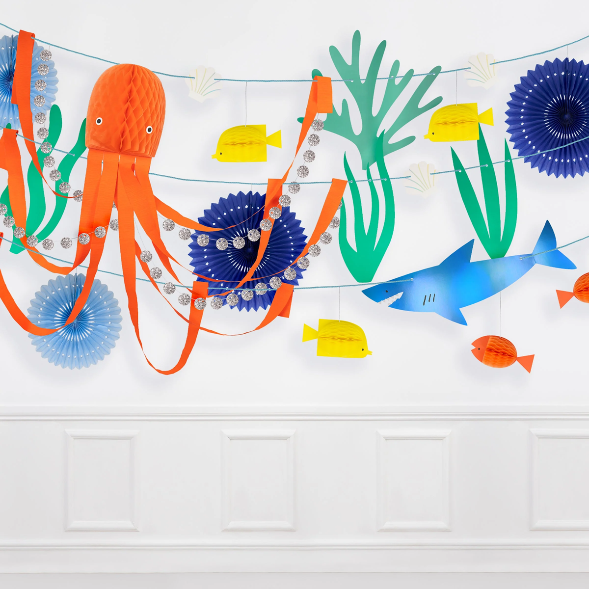 Under the sea garland with paper cutouts of an orange octopus, blue shark, and sea life hanging on a white wall.