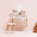 White wooden toy vanity table and stool set with play cosmetics, mirror, and a teddy bear nearby.