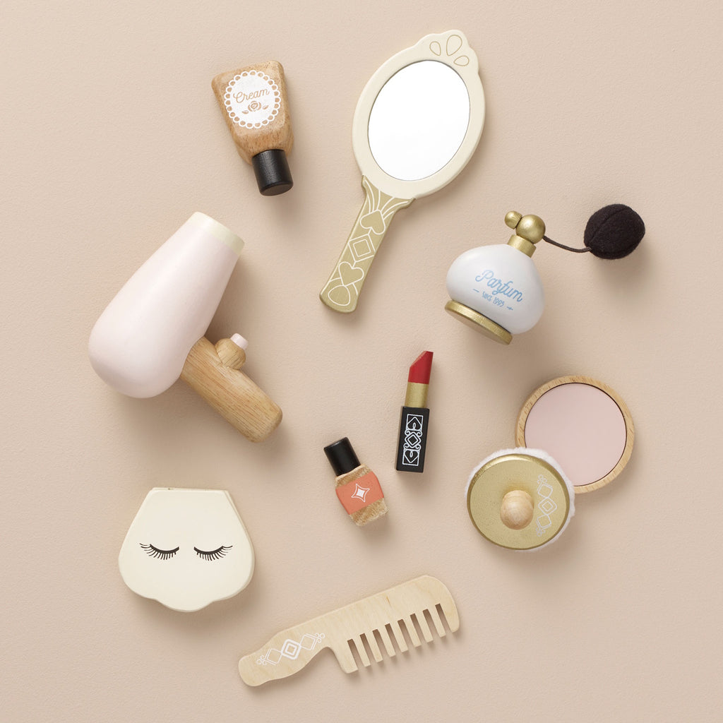 Wooden toy makeup set on a beige background, including a mirror, comb, lipstick, nail polish, hairdryer, cream, perfume, and compact. Playful tone.